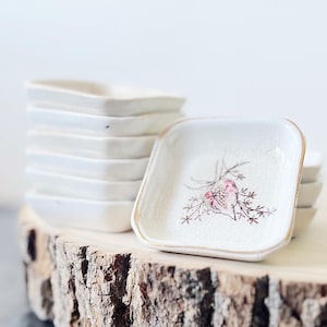 May include: A stack of small, square, white ceramic dishes with a floral design and gold trim. The dishes are stacked on a wooden slab with a natural bark edge. The floral design features pink and brown accents.