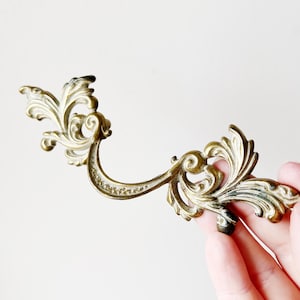 May include: Ornate, antique brass drawer pull with a curved handle and decorative floral scrollwork. The hardware has a vintage aesthetic, with a tarnished finish and intricate details. A classic piece for furniture restoration.