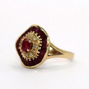Puede incluir: Un anillo de oro con un rubí rojo y un halo de diamantes más pequeños. El anillo tiene un engaste cuadrado.