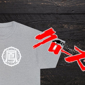 Puede incluir: Una camiseta gris con un gráfico circular blanco con caracteres japoneses. El gráfico está centrado en la camiseta. La camiseta está sobre un fondo de madera negro. El texto "Crows" es visible en rojo en el lado derecho de la imagen.