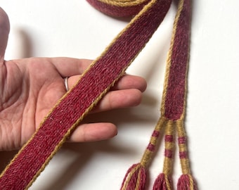 Può includere: Una lunga cintura in tessuto intrecciato caratterizzata da una striscia centrale rosso scuro bordata da profili giallo senape. La cintura è arrotolata a un'estremità e termina con tre fili intrecciati, ciascuno con una nappa di filato rosso e giallo.