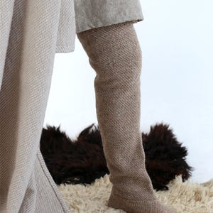 Puede incluir: Un par de botas largas de punto beige con un patrón texturizado. Las botas las lleva una persona de pie sobre una alfombra blanca y esponjosa.