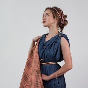 May include: A woman wearing a blue sleeveless tunic with a pink and brown plaid shawl draped over her shoulders. She is wearing a brown belt with a white stripe and a beaded headband.