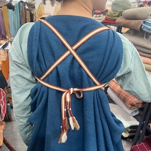 May include: A blue medieval-style dress with a crisscross strap design. The dress has a decorative belt with tassels. The sleeves are a light green color. The dress is made of a thick fabric.