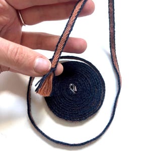 May include: A spool of navy blue and rust-colored woven ribbon. The ribbon is partially unraveled, revealing a coiled section. The ribbon is held by a hand with a silver ring. The ribbon has a textured appearance.