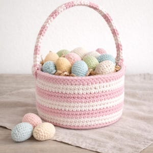 May include: A crocheted Easter basket with a pink and white striped design, filled with colorful crocheted eggs. Several eggs in pastel colors are scattered around the basket on a wooden surface. The basket has a handle.
