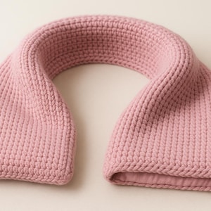 May include: A dusky pink knitted scarf. The scarf is made of a soft, textured material and has a curved shape. The ends are open, and the scarf appears to be hand-knitted. The scarf is a solid colour and is suitable for cold weather.