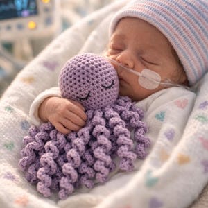 May include: A lavender crochet octopus toy with a round head and curly tentacles, featuring embroidered closed eyes. The toy is held by a newborn infant wearing a white long-sleeved shirt and a light blue and white striped knit cap.