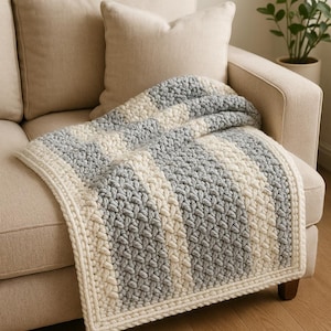 Puede incluir: Una manta tejida a mano en crema, gris y blanco, sobre un sofá beige. La manta presenta un diseño a rayas texturizado. La manta de hilo suave es ideal para añadir calidez y comodidad a un espacio habitable.