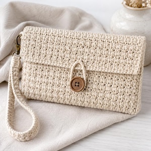 Op de afbeelding: Een rechthoekige, crèmekleurige gehaakte clutch met een gestructureerd steekpatroon, een overslagflap die wordt vastgezet met een enkele bruine houten knoop en een bijpassende afneembare polsband met een bronzen metalen karabijnhaak.