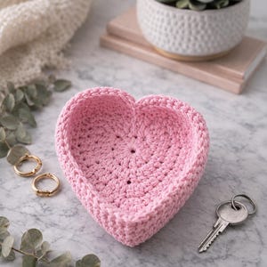 Puede incluir: Una cesta en forma de corazón de ganchillo rosa claro. La cesta está sobre una superficie de mármol, con dos anillos dorados y una llave. La cesta probablemente se utiliza para guardar cosas o para decoración.