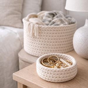 Puede incluir: Dos cestas de ganchillo color crema sobre una superficie de madera. La cesta más pequeña contiene anillos y joyas doradas. La cesta más grande está llena de una manta doblada. También se ven una lámpara y un jarrón decorativo.