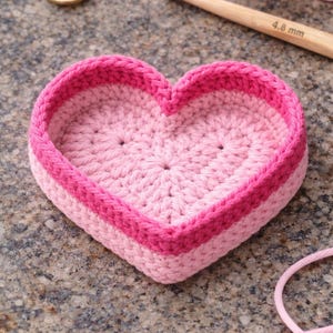 May include: A heart-shaped crochet basket in pink hues. The basket has a light pink interior and a darker pink border. A crochet hook and scissors are in the background. The hook is 4.8 mm.