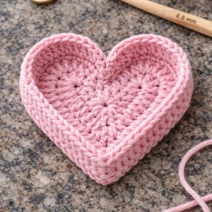 May include: A handmade, pink crochet heart-shaped basket. The basket is made with a textured stitch and has a raised border. A wooden crochet hook, measuring 4.8 mm, and gold scissors are visible in the background, along with a ball of pink yarn.