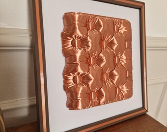 May include: Framed artwork with a copper-coloured, padded square with a buttoned design. The frame is brown with an orange stripe, set against a white background. The artwork is displayed on a wooden surface.