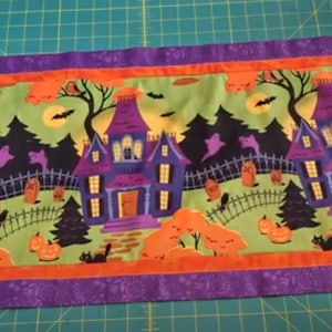 May include: A Halloween table runner with a green background featuring a repeating pattern of purple houses with orange pumpkins, black cats, and bats. The runner has a purple, orange, and white border.