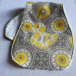 May include: A grey and yellow baby bib with a yellow rubber ducky appliqué. The bib has a white terry cloth backing.