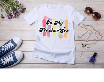 Może przedstawiać: Biała koszulka z napisem "In My Teacher Era" w stylu retro, ozdobiona kolorowymi pionowymi paskami i kwiatami. Na zdjęciu znajdują się również niebieskie trampki, okulary przeciwsłoneczne i biżuteria, ułożone na jasnej drewnianej powierzchni.