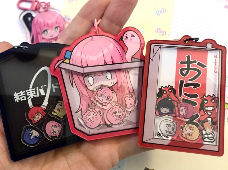 May include: A set of six colorful acrylic keychains featuring anime characters. The keychains are shaped like squares and rectangles with clear plastic windows. The characters are depicted in a chibi style with large heads and small bodies. The keychains are attached to metal rings.
