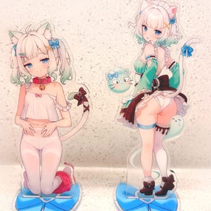 Puede incluir: Dos figuras de acrílico con personajes estilo anime con orejas y colas de gato. La figura de la izquierda está arrodillada con un top blanco y medias con un collar rojo. La figura de la derecha viste un conjunto verde y marrón con un lazo azul.