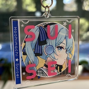 May include: A clear acrylic keychain featuring a cartoon character with blue hair and a pink bow. The character is surrounded by a circle with the word "SUISEI" written in pink letters. The text "Hoshimachi Suisei" is written vertically on the left side of the keychain.