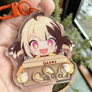 May include: A clear acrylic keychain featuring an anime-style character with blonde and brown hair, wearing a yellow jacket and holding a box with the word "Grems" on it. The character has bright red eyes and a wide smile. Inside the box are four small, cartoon-like fish.