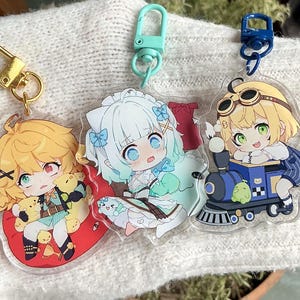 May include: Three anime character keychains with different designs. The first keychain features a yellow-haired character with a red and white outfit sitting on a red cushion. The second keychain features a blue-haired character with a white and blue outfit sitting on a white cushion. The third keychain features a yellow-haired character with a blue and white outfit sitting on a blue train.