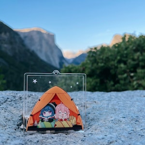 May include: A clear acrylic keychain featuring two anime characters inside an orange tent with stars in the background.