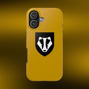 May include: A mustard yellow smartphone case featuring a centered black shield emblem with a white stylized badger face graphic. The case has a dual-lens camera cutout in the upper left corner and a smooth, solid-colored finish on the back surface.