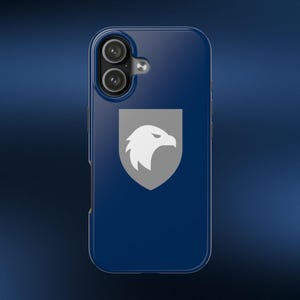May include: A dark blue smartphone case featuring a centered grey shield emblem with a white eagle head silhouette. The case has a dual-camera cutout at the top left with a small circular flash opening, maintaining a smooth, glossy finish throughout.