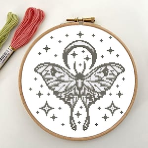 Peut inclure: Un motif de point de croix noir et blanc représentant un papillon de nuit avec une lune croissante et des étoiles sur fond blanc. Le motif est encadré dans un cercle en bois.