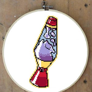 May include: A pixelated cross-stitch design of a lava lamp in a wooden embroidery hoop. The lamp features a red top and base, a yellow midsection, and a light blue and purple interior. The design is set against a white background.