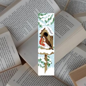May include: A pixel art bookmark featuring a robin perched on a birdhouse in a snowy winter scene. The birdhouse is brown with a white roof and a small opening. The robin is red, white, and brown. The background is white with blue and green accents.