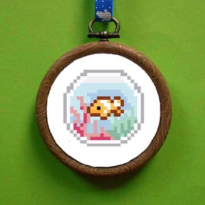 May include: A wooden framed embroidery hoop with a white background and a pixelated image of an orange and white clownfish swimming in a blue and white fishbowl with pink coral and green seaweed.