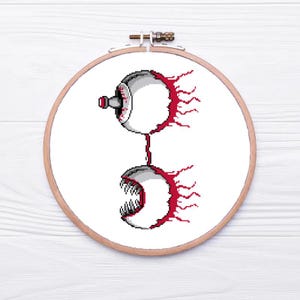 May include: A pixel art cross-stitch design of two stylized eyes in a wooden embroidery hoop. The top eye is gray and white with a red accent, connected to a lower eye with sharp teeth. The design is on a white background.