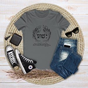 Puede incluir: Una camiseta gris con un dise&ntilde;o gr&aacute;fico negro y texto. El dise&ntilde;o incluye texto hebreo y una corona de trigo. La camiseta se combina con vaqueros azules, zapatillas altas negras, una cartera negra y gafas de sol sobre una estera tejida.