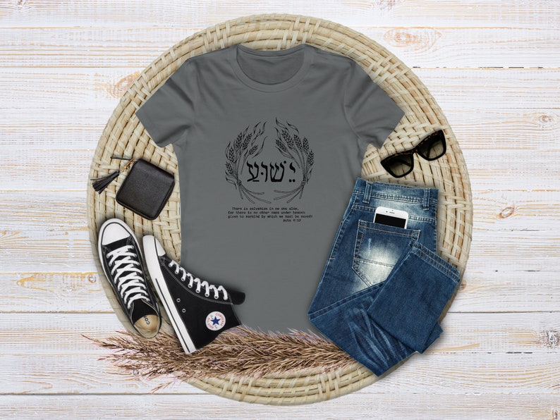 Puede incluir: Una camiseta gris con un dise&ntilde;o gr&aacute;fico negro y texto. El dise&ntilde;o incluye texto hebreo y una corona de trigo. La camiseta se combina con vaqueros azules, zapatillas altas negras, una cartera negra y gafas de sol sobre una estera tejida.
