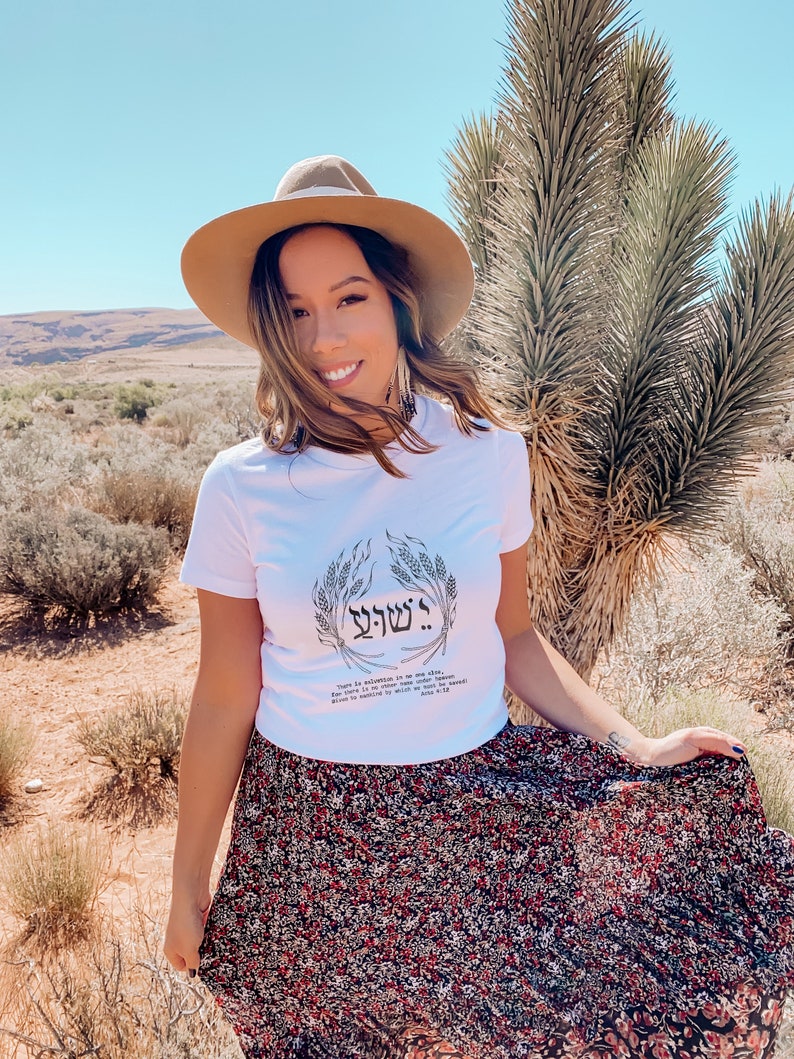 Puede incluir: Una mujer con una camiseta blanca con texto hebreo y un dise&ntilde;o de trigo, combinada con una falda floral y un sombrero marr&oacute;n. La imagen est&aacute; tomada al aire libre con un paisaje des&eacute;rtico de fondo.