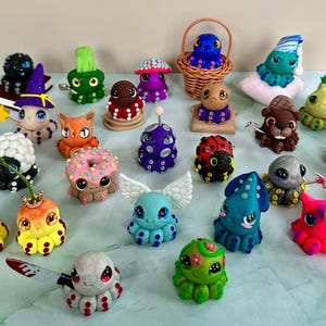 Puede incluir: Una colección de figuras de pulpo de arcilla caprichosas y coloridas, cada una con diseños y accesorios únicos. Algunos pulpos usan sombreros, tienen alas o sostienen objetos como un cuchillo o una varita. Las figuras están dispuestas sobre una superficie azul claro.