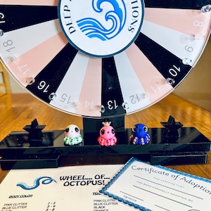 May include: A colorful octopus-themed prize wheel with three miniature octopus figurines. The wheel has a Deep Sea Clay Creations logo and is surrounded by a black and white striped pattern. A certificate of adoption and a color chart are also visible.