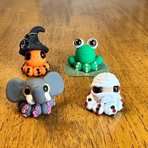May include: Four whimsical clay figurines: an orange octopus wearing a witch hat, a green frog on a leaf, a gray elephant, and a white mummy. Each figure has large, expressive eyes and colorful details.