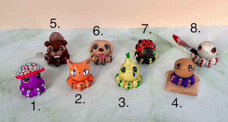 Pu&ograve; includere: Una collezione di otto figurine di argilla fatte a mano. Le figure includono un polpo, un gatto, una cipolla, un cane, un castoro, una coccinella e un polpo con un coltello. Ogni figura &egrave; dai colori vivaci e dettagliata, che mostra un design unico.
