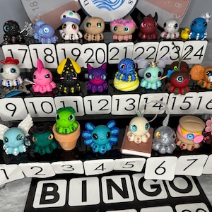Pode incluir: Uma coleção de pequenas e coloridas figuras de polvo esculpidas, exibidas num suporte preto de vários níveis numerado de 1 a 24. As figuras apresentam vários temas, incluindo animais, comida e personagens de fantasia, com cores distintas.