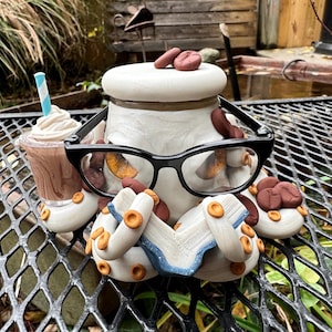 Pode incluir: Uma figura de polvo de cerâmica branca usando óculos e segurando um livro. O polvo tem grãos de café marrons na cabeça e nos tentáculos. O polvo está segurando uma xícara de café com um canudo.