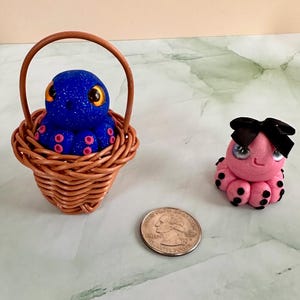 Pu&ograve; includere: Figurine di polpo. Un polpo blu glitterato in un piccolo cestino di vimini, accanto a un polpo rosa con un fiocco nero. Il polpo blu ha ventose rosa e occhi gialli. Accanto c'&egrave; una moneta.