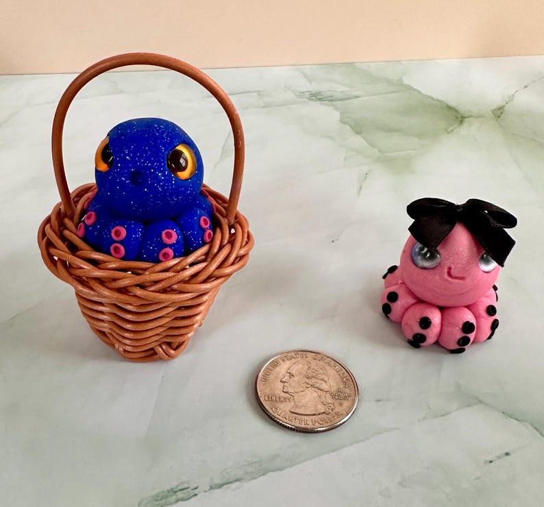 Pu&ograve; includere: Figurine di polpo. Un polpo blu glitterato in un piccolo cestino di vimini, accanto a un polpo rosa con un fiocco nero. Il polpo blu ha ventose rosa e occhi gialli. Accanto c'&egrave; una moneta.
