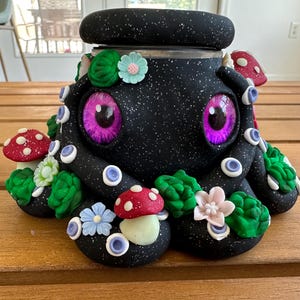 Könnte beinhalten: Ein schwarzes Gefäß mit gesprenkelter Oberfläche in Form eines Oktopus mit großen, leuchtend violetten Augen. Die Außenseite ist mit kleinen rot-weißen Pilzen, grünen sukkulentenartigen Clustern sowie hellblauen, rosa und weißen Blüten verziert.