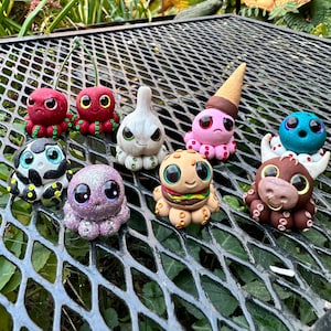 May include: A collection of eight handmade octopus figurines, each with unique details and colors. The octopuses are made of polymer clay and have different expressions and features, including a burger, a cherry, a garlic, and a cow.
