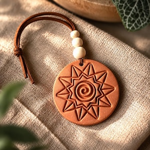 May include: A round terracotta-colored clay ornament featuring an embossed sun design with a central spiral. The ornament is attached to a brown leather cord accented with three small, round, light-colored wooden beads.