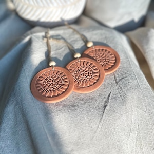 May include: Three terracotta-colored, circular ornaments with a sunflower design are displayed on a light gray fabric. Each ornament has a jute string and a wooden bead for hanging. The ornaments are likely for home decor.